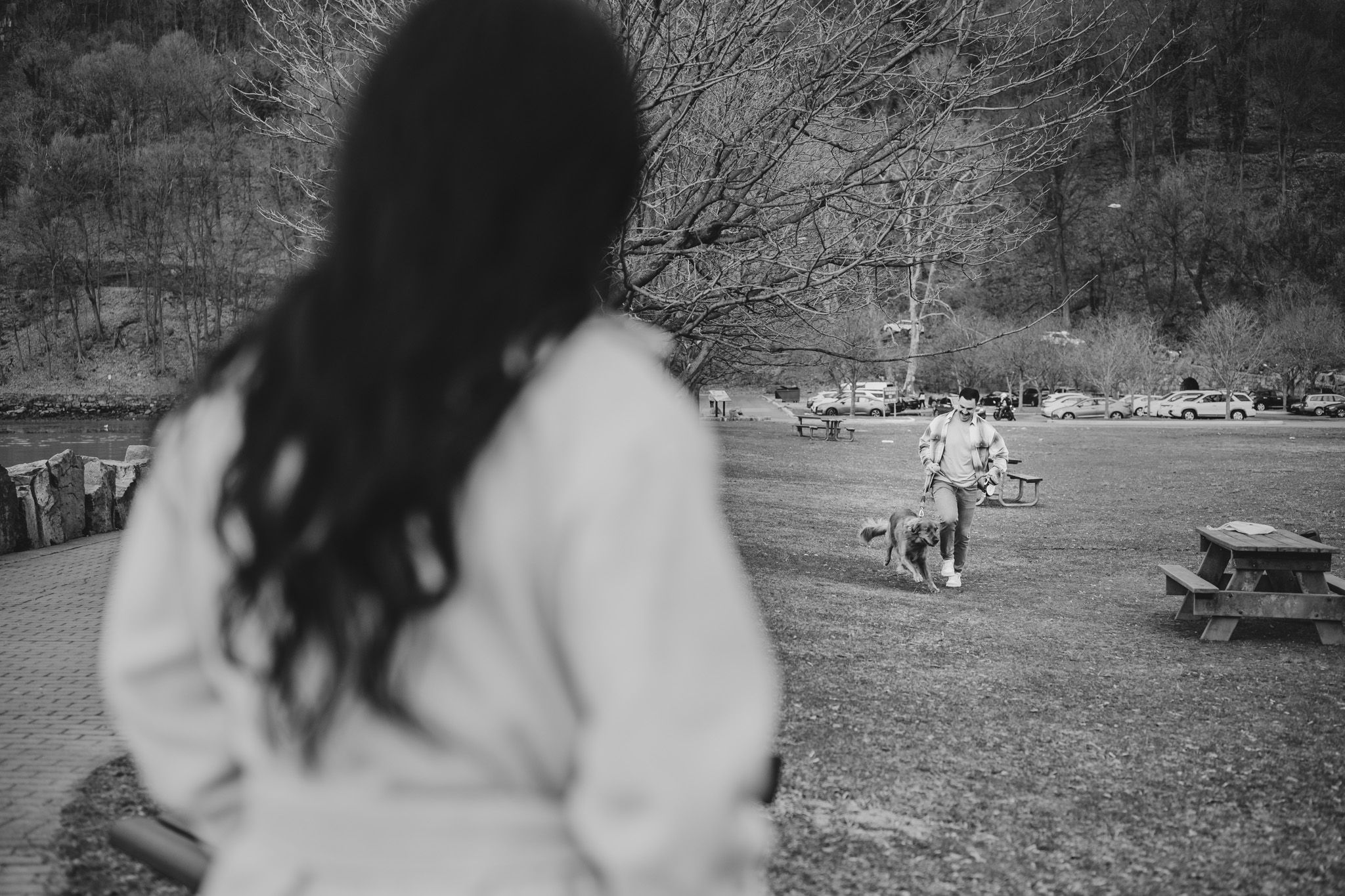 Black and white photo of a woman with long dark hair watching a person walk a dog in a park.