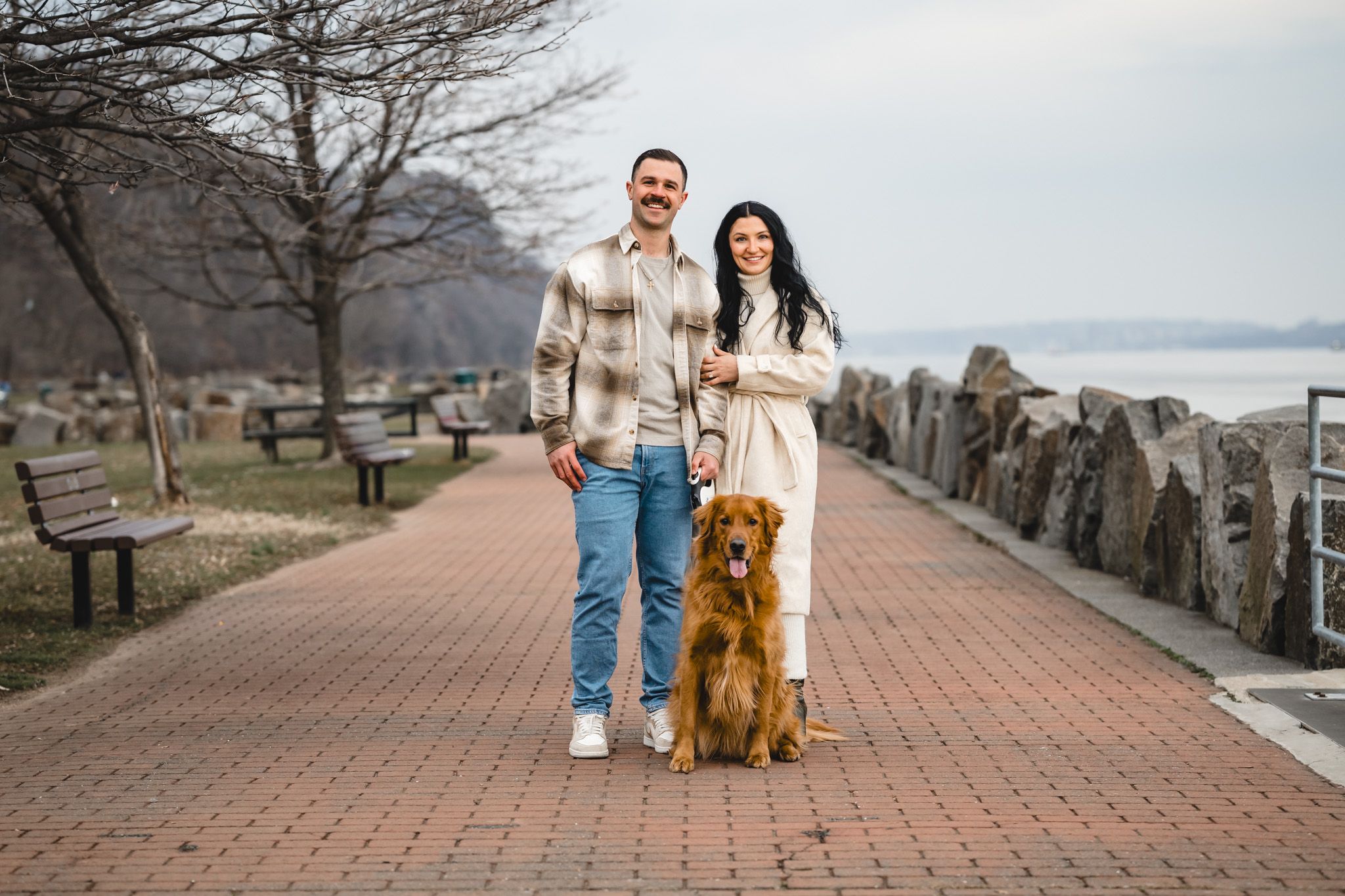 Couple with golden retriever dog posing on brick walkway near waterfront park with rocky shoreline.