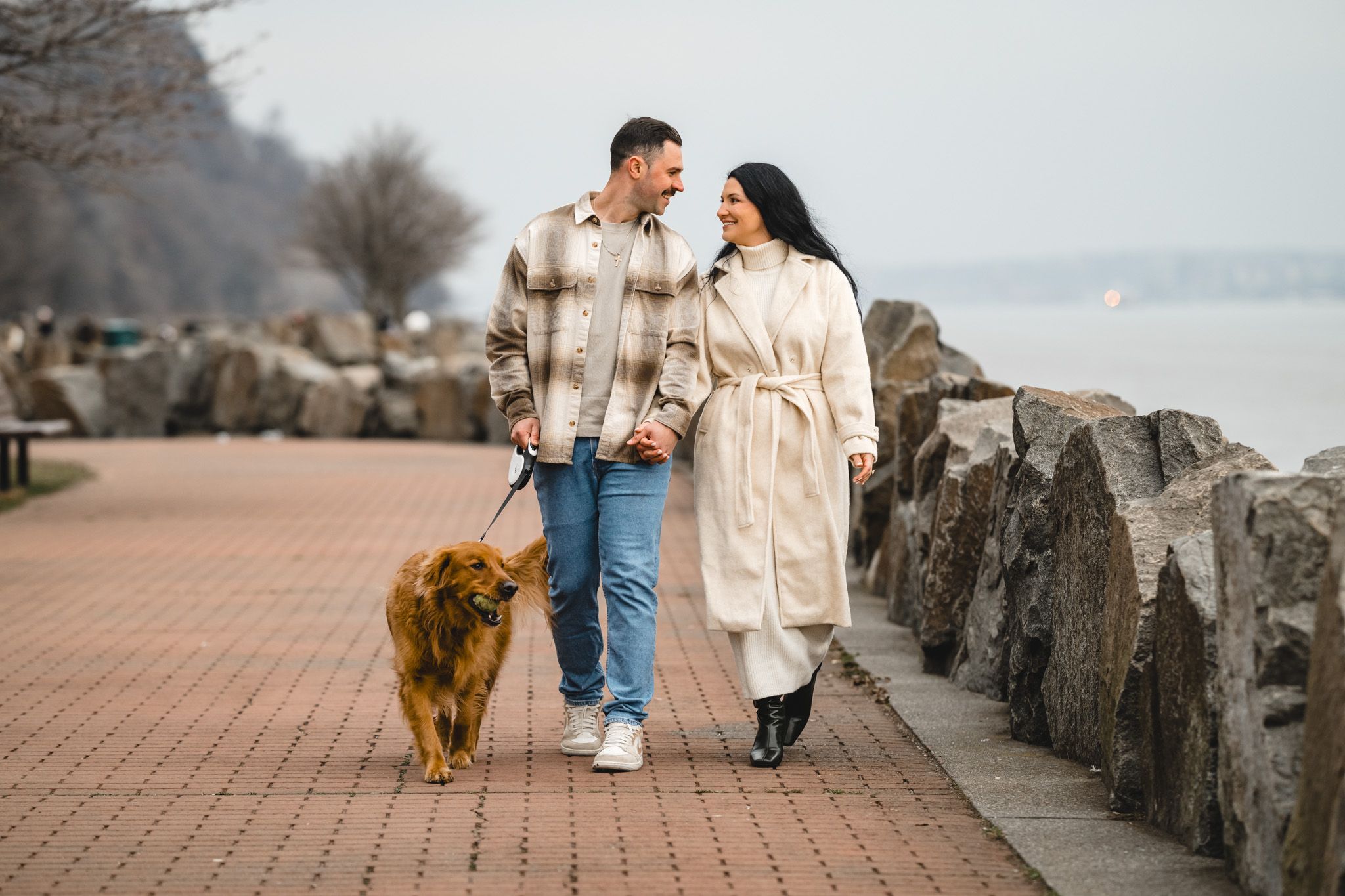 Couple walking golden retriever dog on waterfront promenade in autumn, holding hands and smiling at each other.