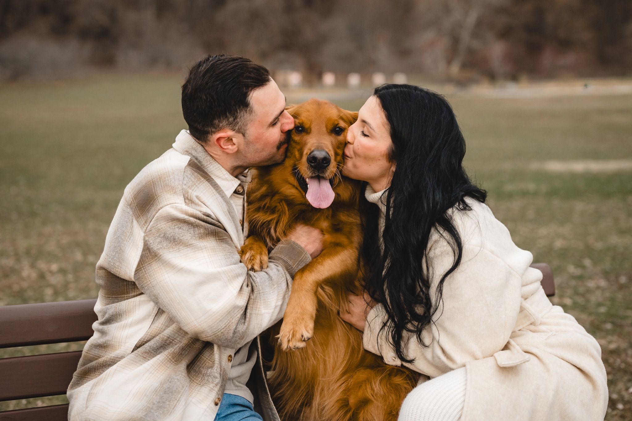 Happy couple kissing their golden retriever dog on a park bench during an outdoor autumn photo session.