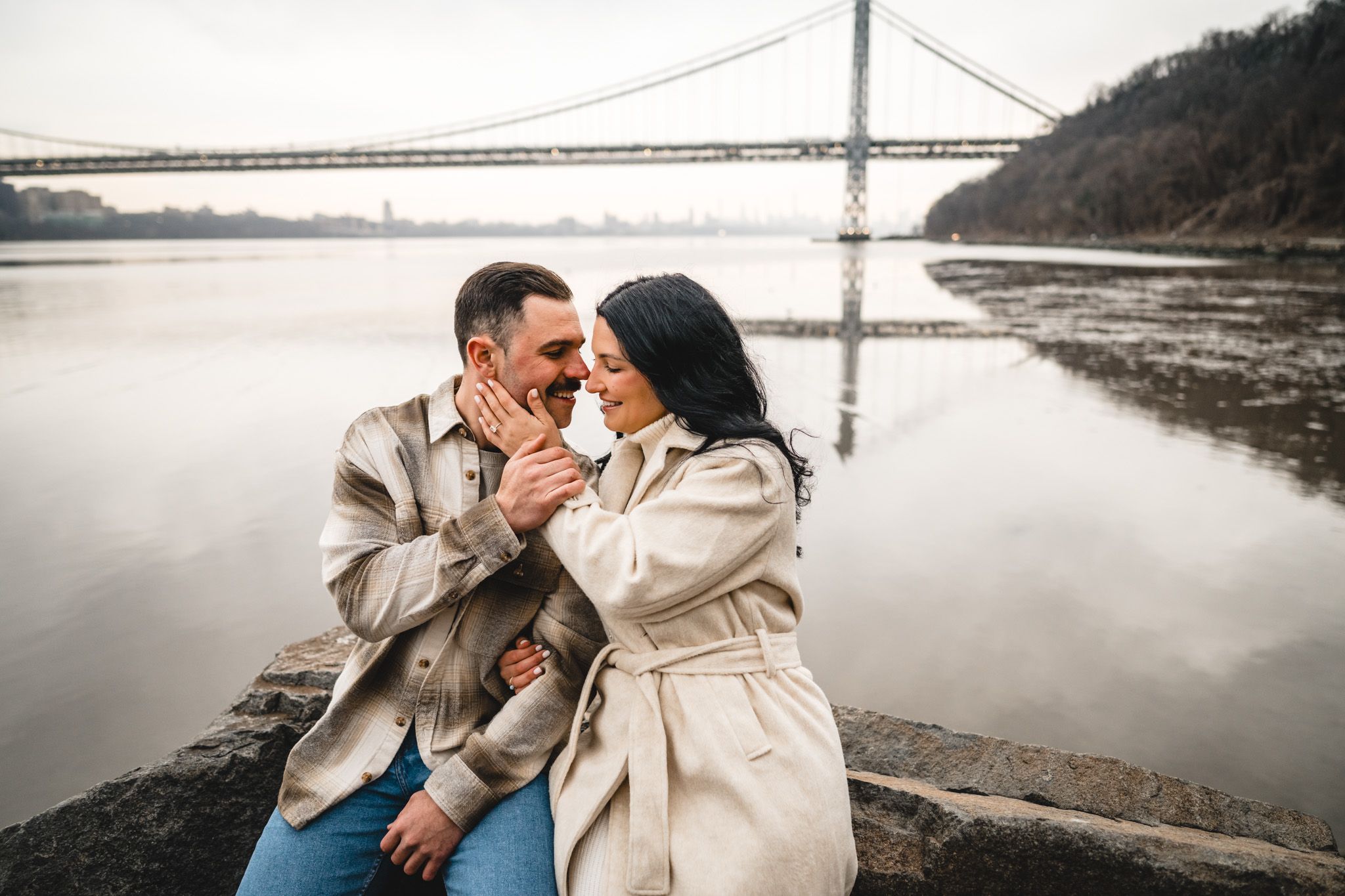 Romantic couple sitting by a river with a bridge backdrop on a cloudy day, sharing a tender moment.