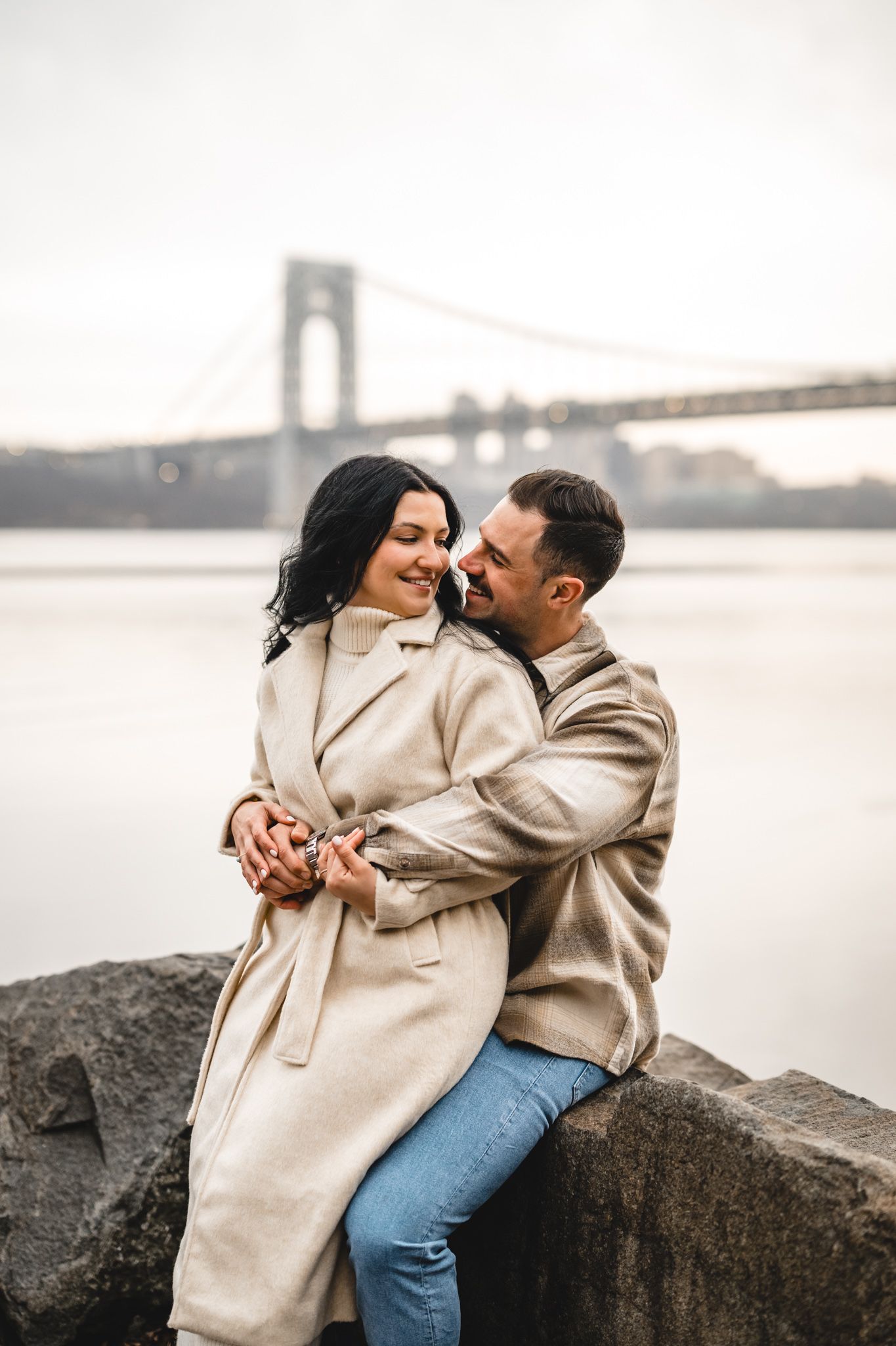 Romantic couple embracing near waterfront with iconic suspension bridge in background on cloudy day.