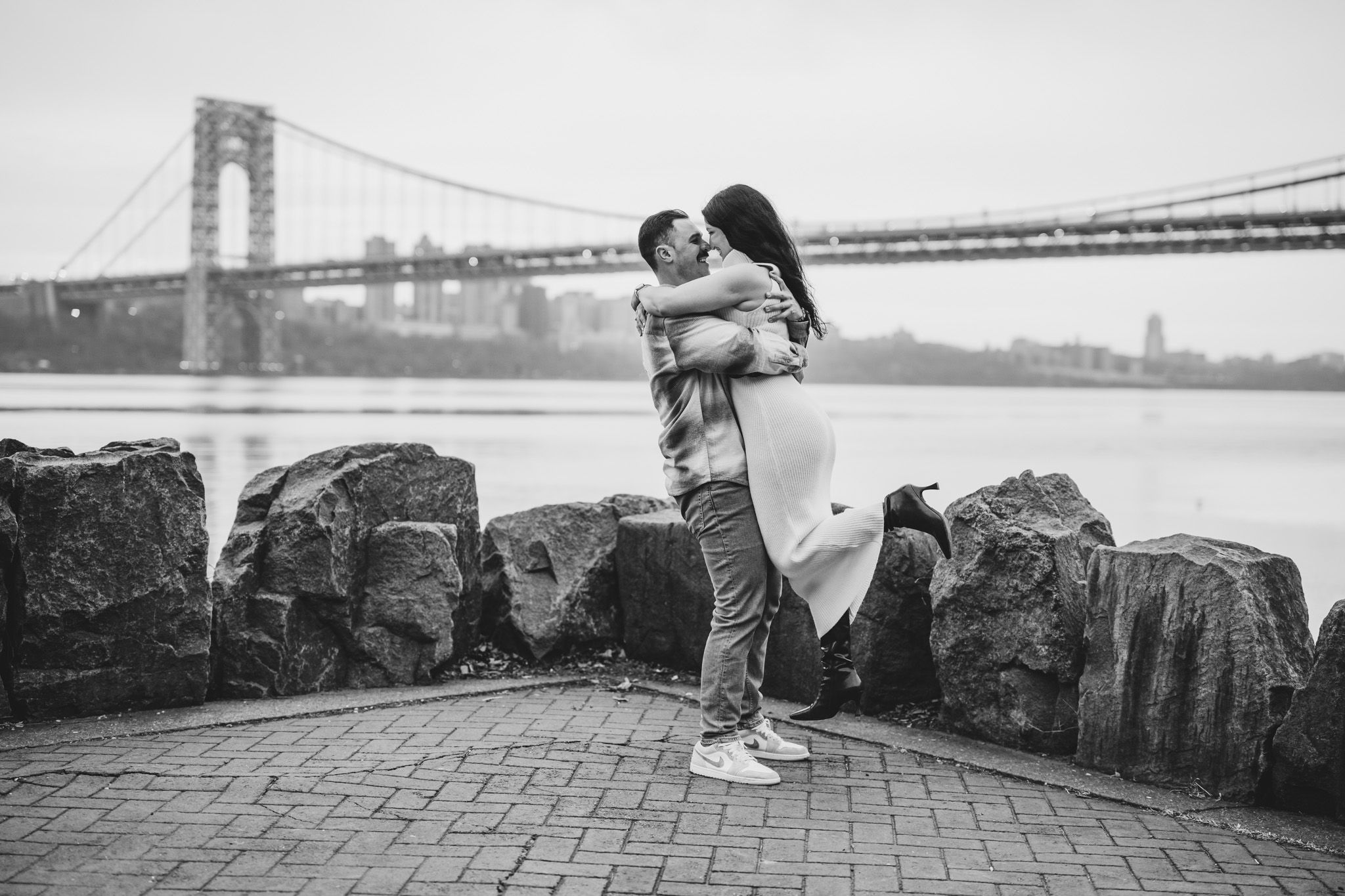 Black and white photo of a couple embracing near rocky waterfront with George Washington Bridge in background.