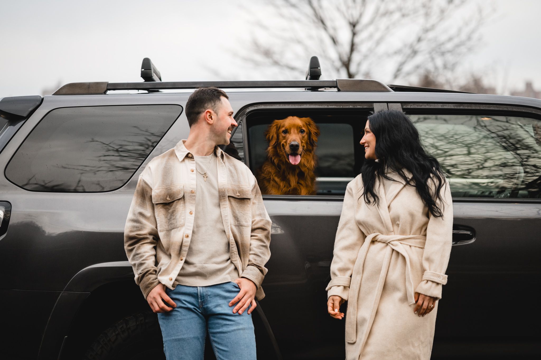 Couple smiling at each other beside a black SUV with a golden retriever dog looking out the rear window.