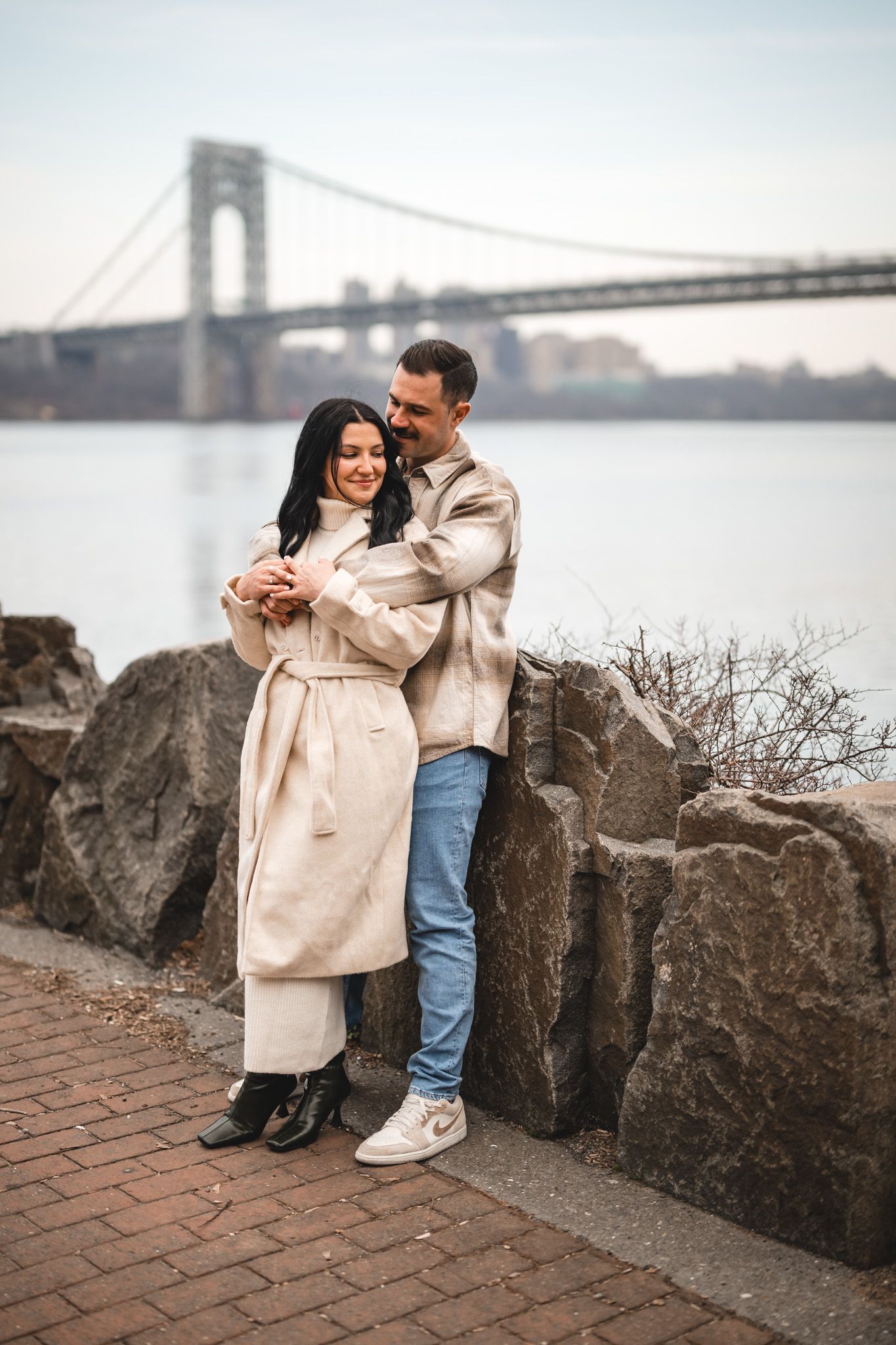 A couple embraces near rocky waterfront with George Washington Bridge in background on a cloudy day.