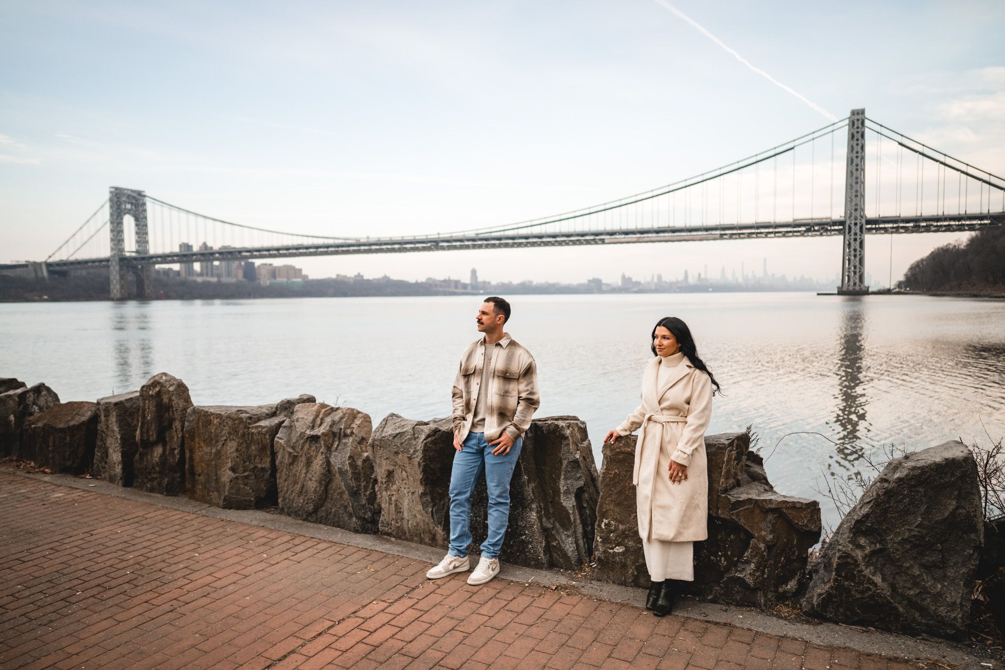 Couple standing apart near rocky waterfront with George Washington Bridge spanning river in background on overcast day.