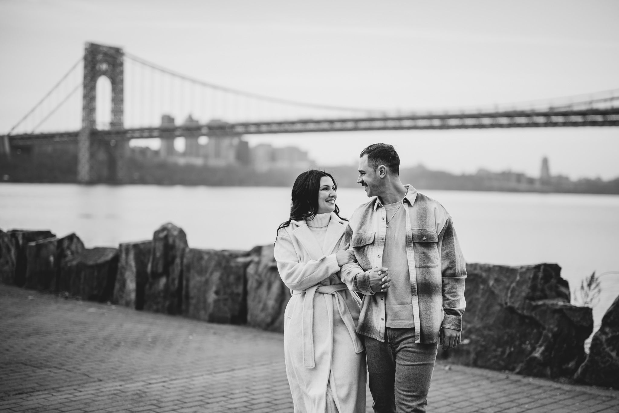 Black and white photo of a couple smiling at each other by the George Washington Bridge on the Hudson River.