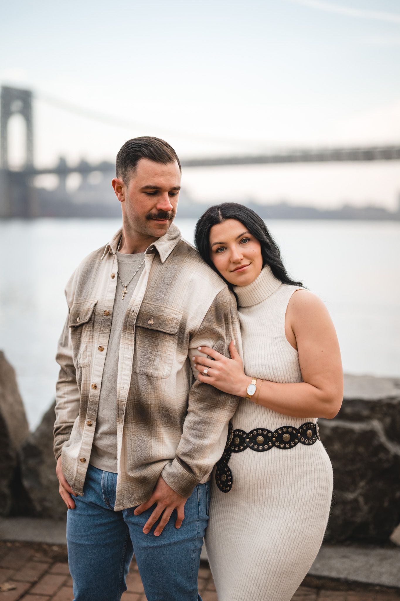 Couple posing together near a waterfront with a suspension bridge in the background at dusk.