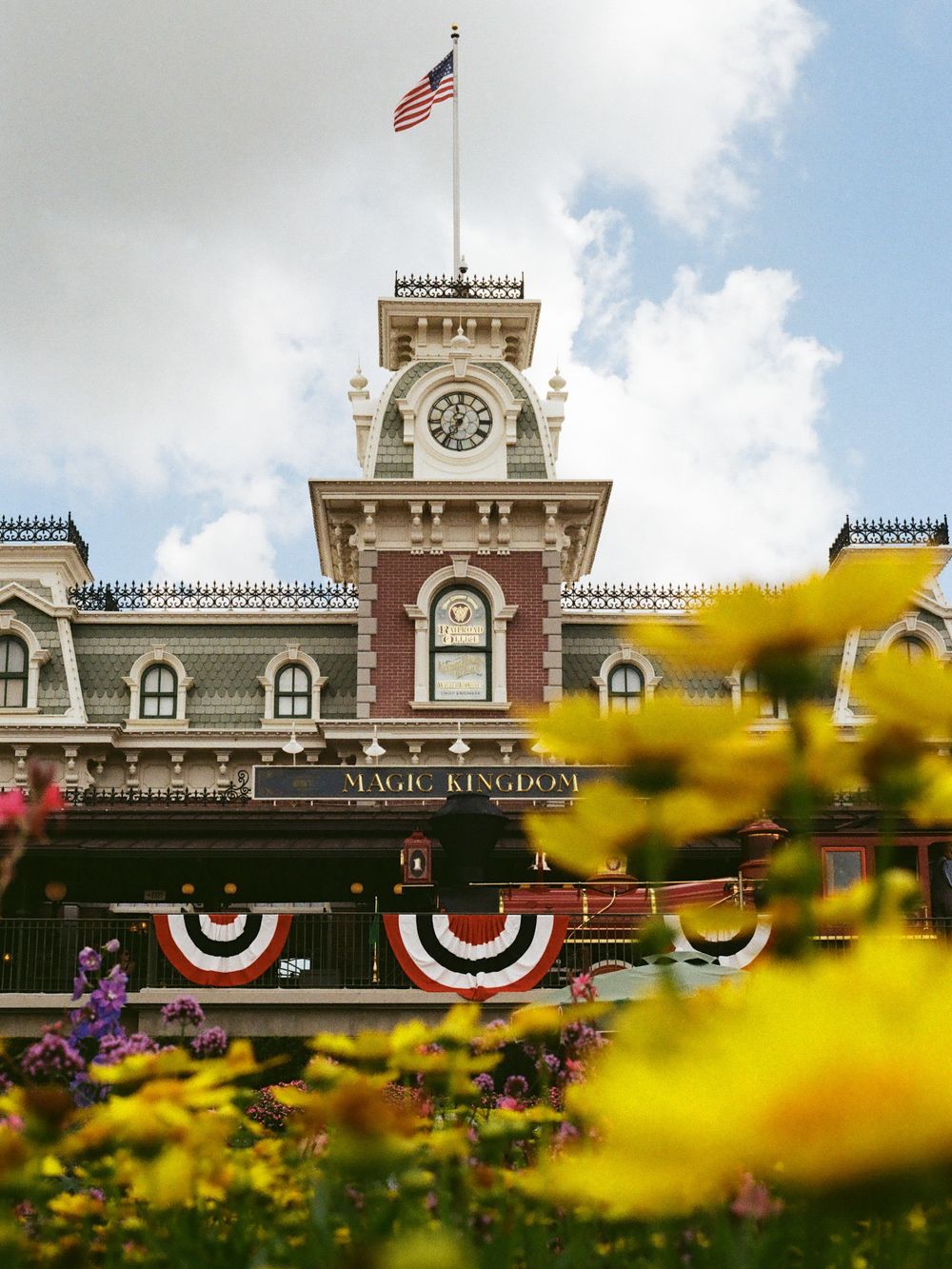 Magic Kingdom train station entrance with American flag, clock tower, and patriotic bunting framed by yellow flowers.