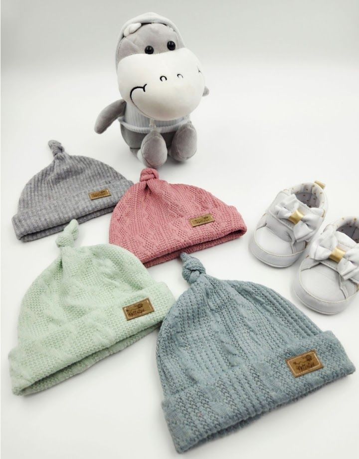 Four colorful knit baby beanies in gray, pink, mint, and blue displayed with a hippo plush toy and white baby sneakers.