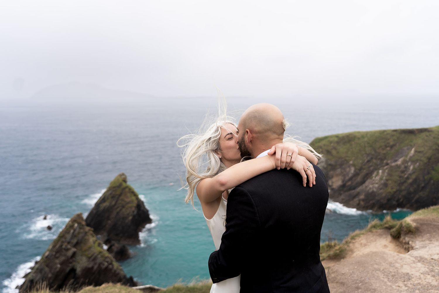 A couple embraces while standing on a scenic coastal cliff overlooking the turquoise ocean and rugged shoreline.