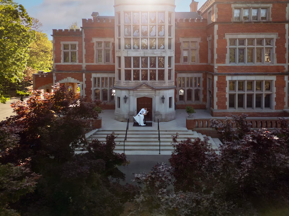 Aerial view of a grand red brick Victorian building with a figure in white on the front steps, surrounded by lush trees.