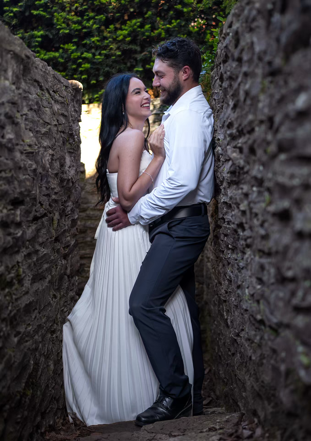 Bride and groom embracing between stone walls, smiling joyfully in wedding attire outdoors.