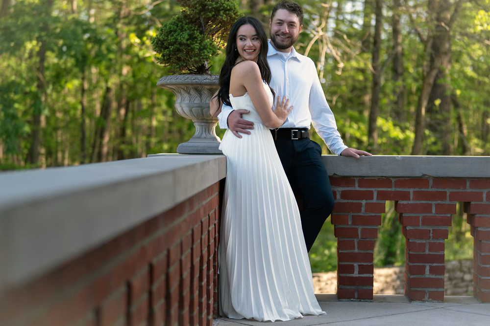 Couple posing on brick bridge in lush garden, woman in white pleated gown, man in blue shirt and dark pants.