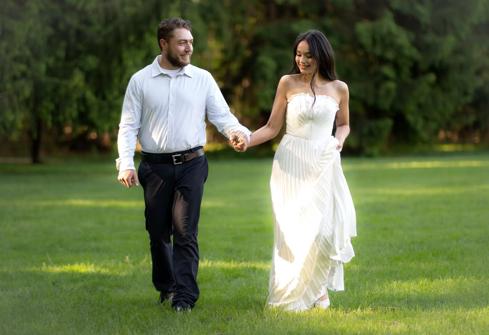 Couple in wedding attire holding hands while walking on green grass with trees in background.