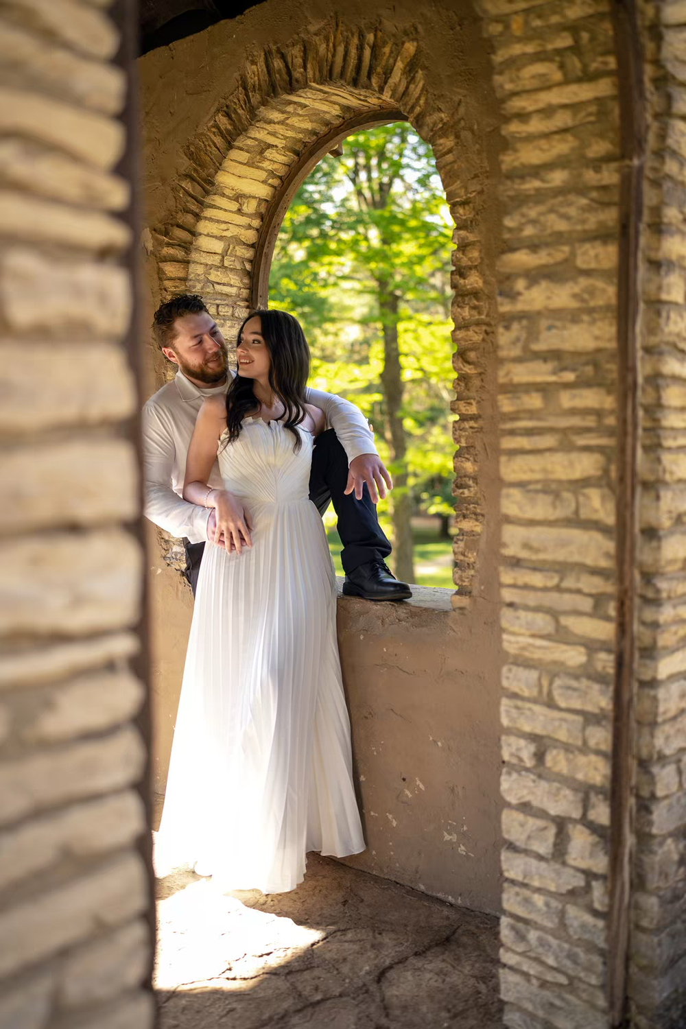 A couple in wedding attire embraces in a stone archway surrounded by lush green trees on their wedding day.