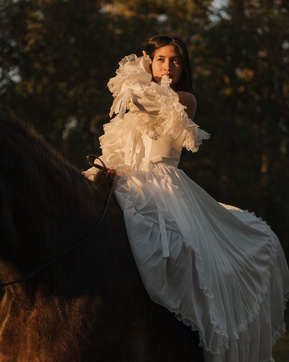 Woman in white ruffled gown riding a dark horse at golden hour with lush green trees in background during photoshoot in sydney with a horse