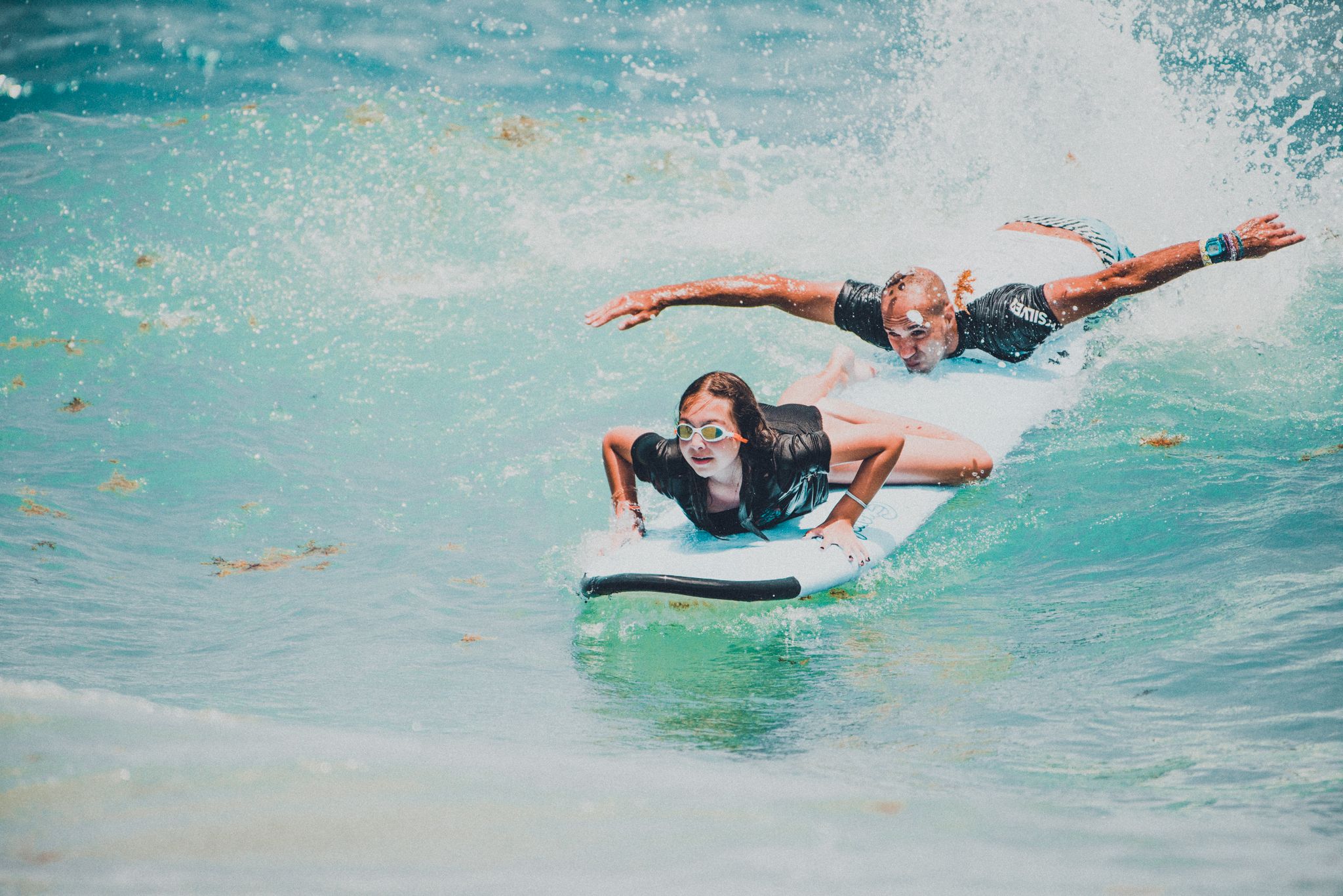 Two surfers riding waves on surfboards in turquoise ocean water, one lying prone and one crouching during an exciting surf session.