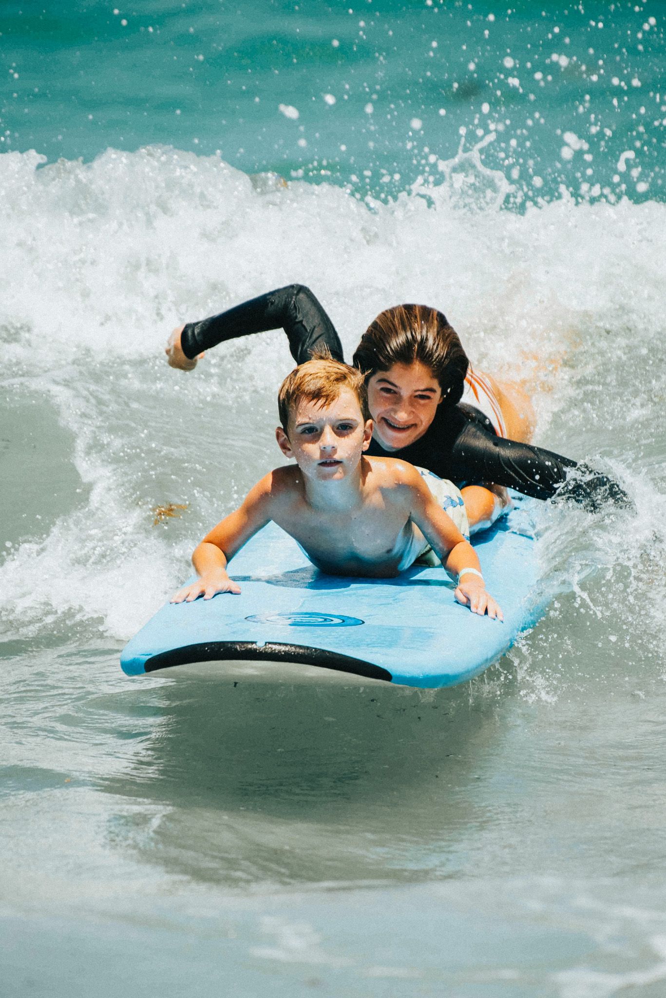 Two kids riding a blue boogie board together through ocean waves on a sunny beach day.