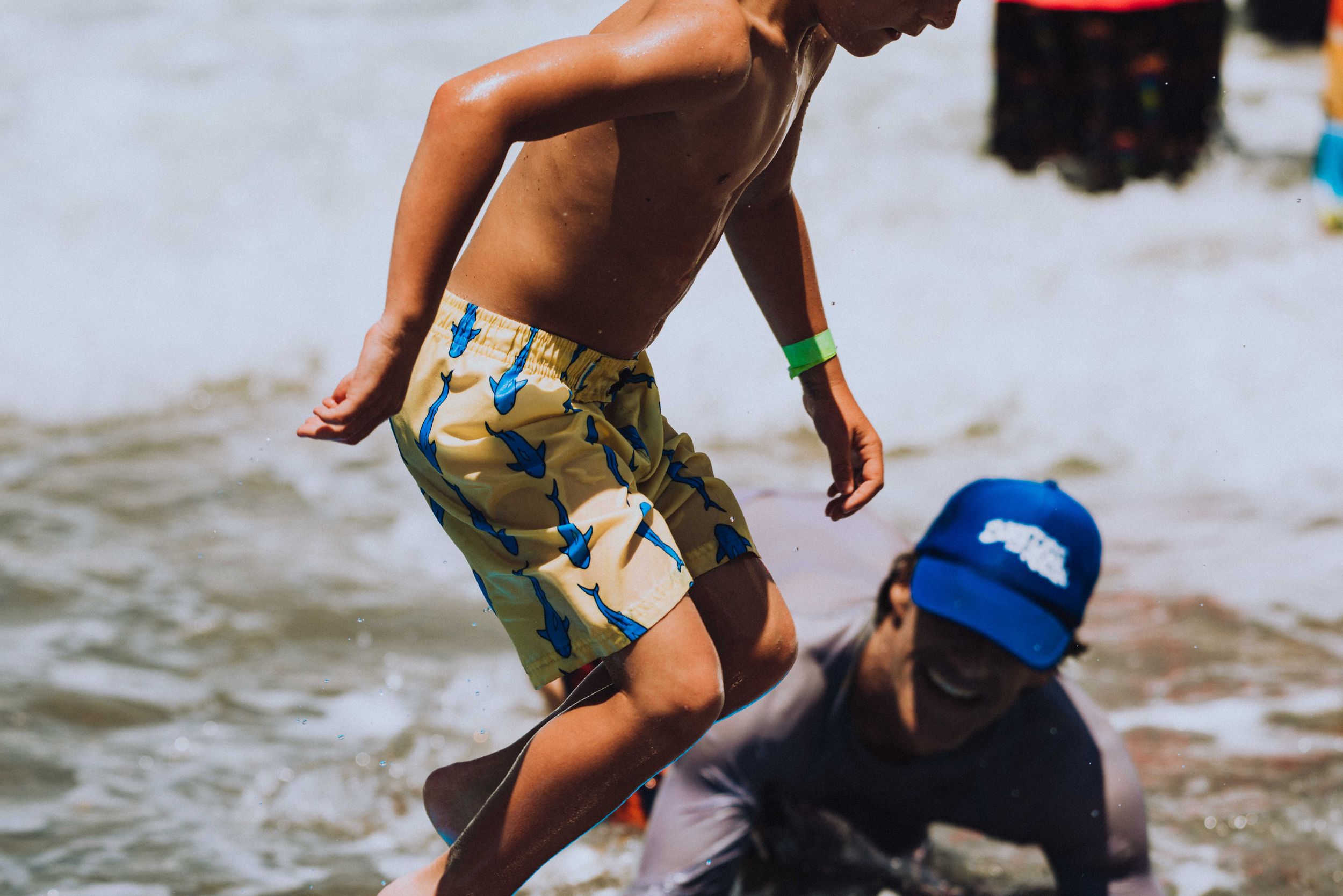 Two children playing in ocean waves, one boy wearing yellow swim trunks and a green wristband at a beach event.