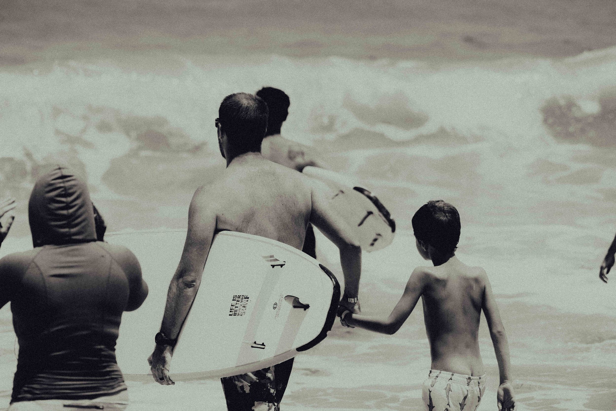 Black and white photo of surfers, including a child, carrying surfboards into ocean waves on a beach.