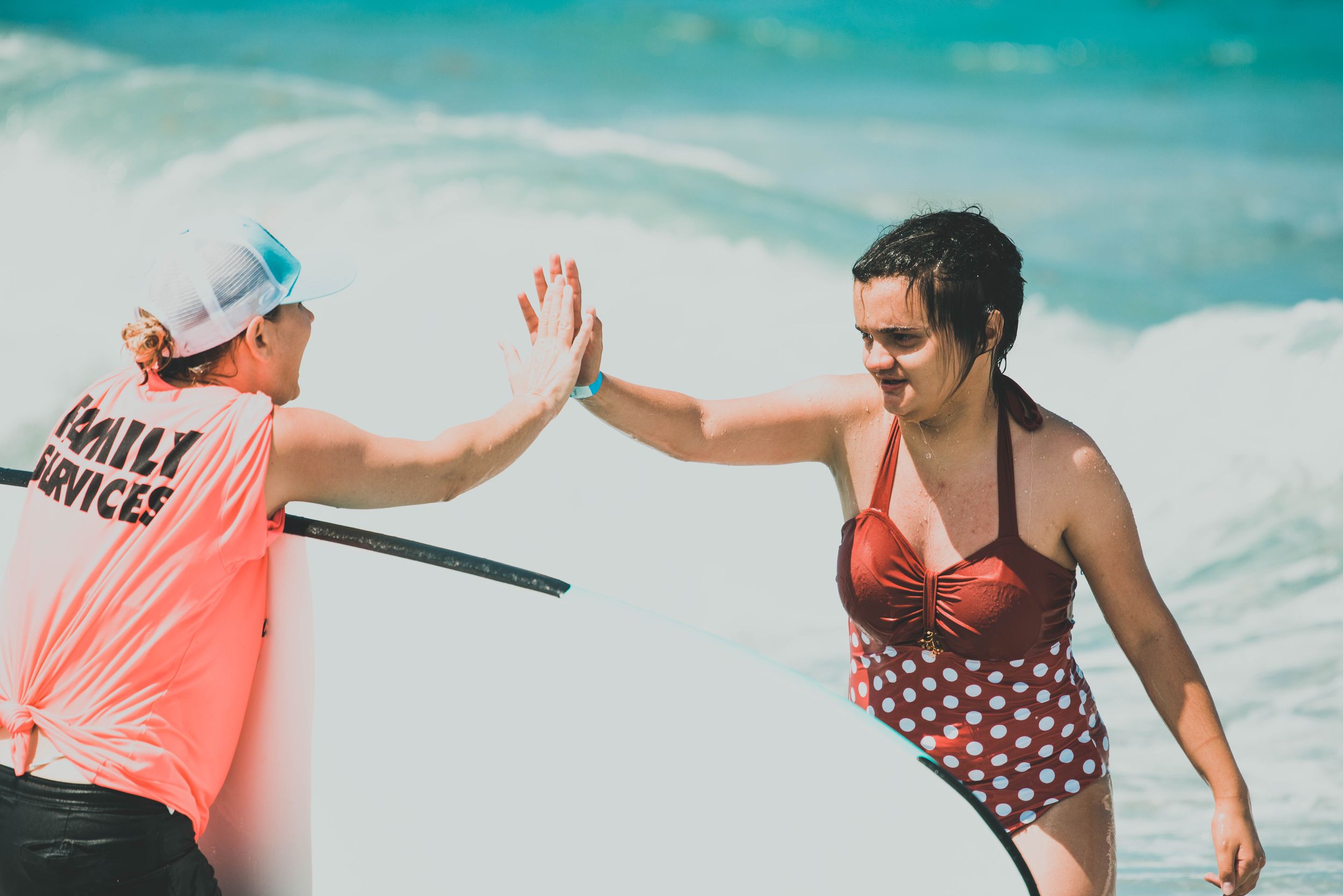 Two women high-fiving on a sunny beach, one wearing a coral Family Services shirt and cap, the other in a red polka dot swimsuit.