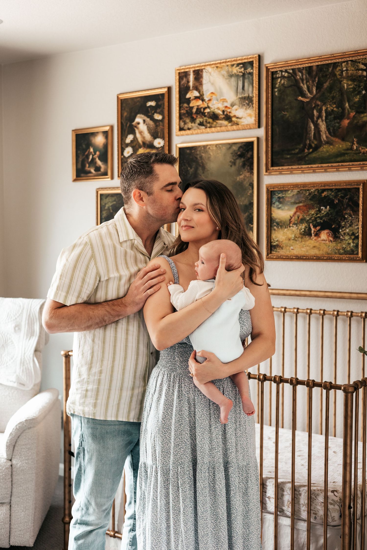 A couple with a newborn baby stands in a nursery with gold-framed nature paintings  | Newborn Photographer in Broomfield Colorado