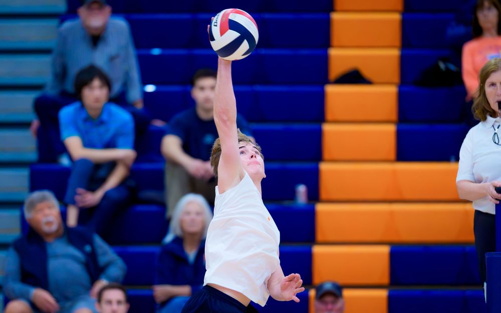Hershey Trojan reaches up to serve the volleyball with fans visible in the stands by Deanna Fox, SportsFox Photography based in Hershey PA.
