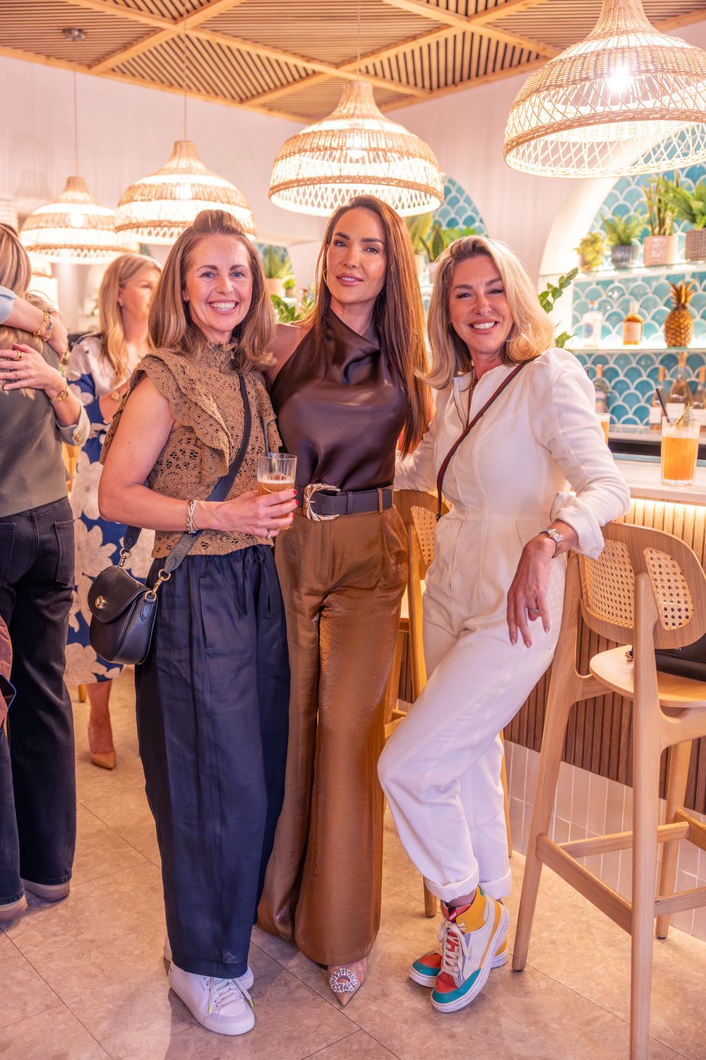 Three stylish women pose together smiling in a modern restaurant with rattan pendant lights.