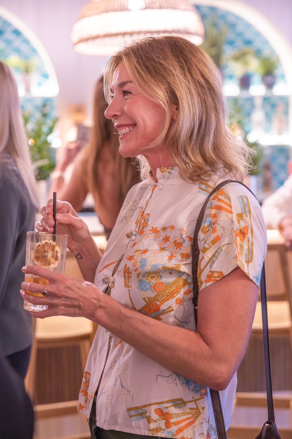 Smiling blonde woman holding a cocktail drink with a straw at a social gathering in a stylish bar.