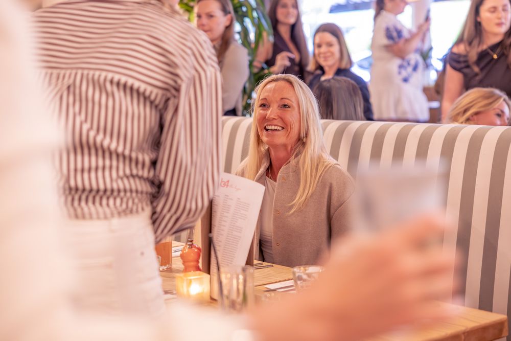 A smiling blonde woman sits in a striped booth at a bright restaurant reviewing a menu.
