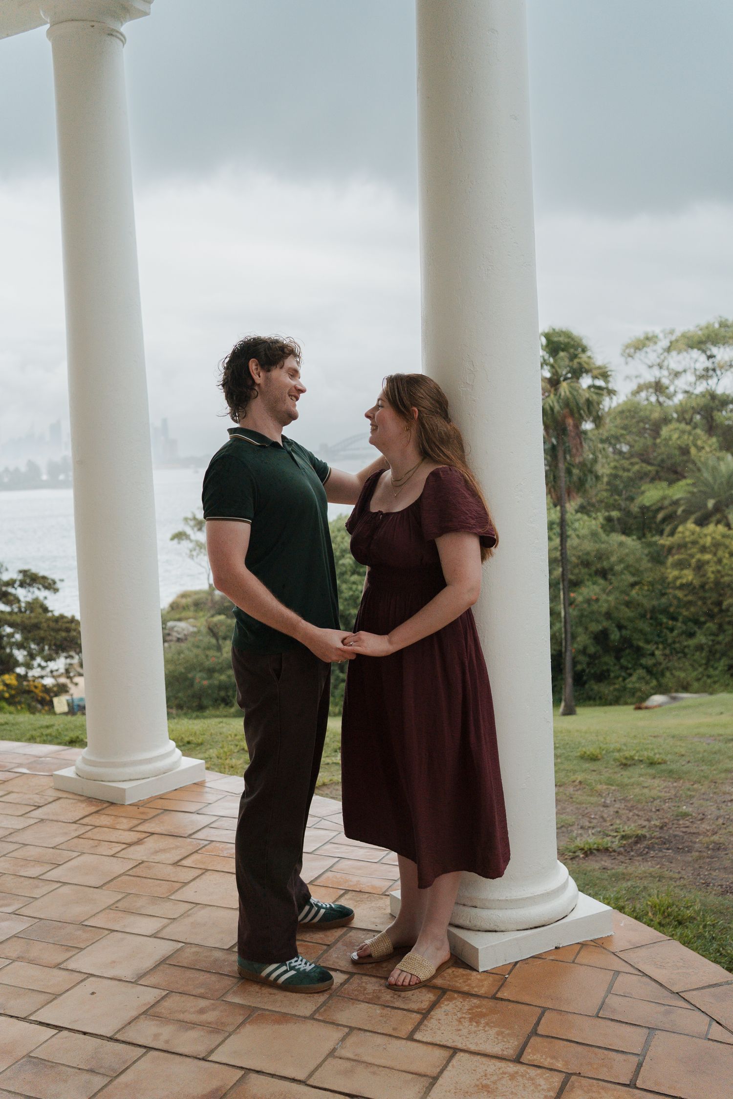 Couple holding hands and smiling at each other between white columns overlooking a waterfront on a cloudy day engagement photoshoot