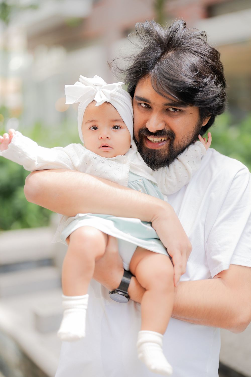 Smiling bearded man holding a cute baby girl in a white bow headband and dress outdoors.