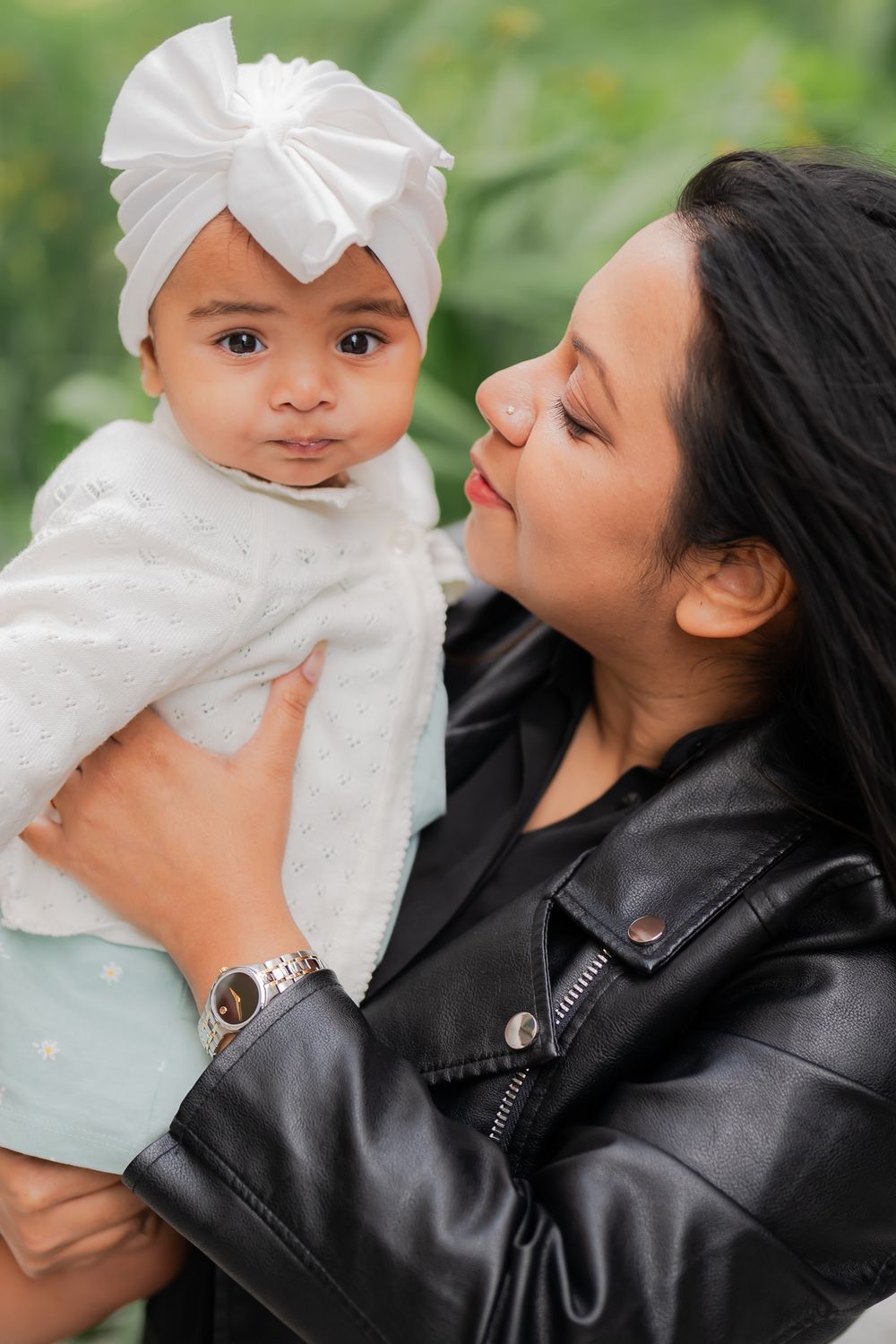A woman in a black leather jacket lovingly holds a baby wearing a white turban bow, outdoors with green foliage.