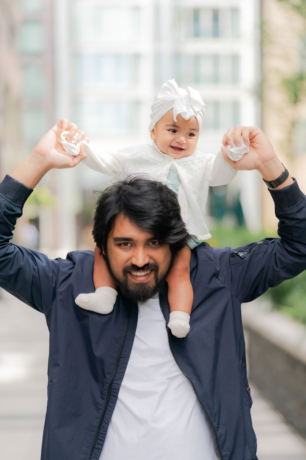 Smiling bearded father holds baby girl on his shoulders outdoors, baby wearing white dress and bow headband.