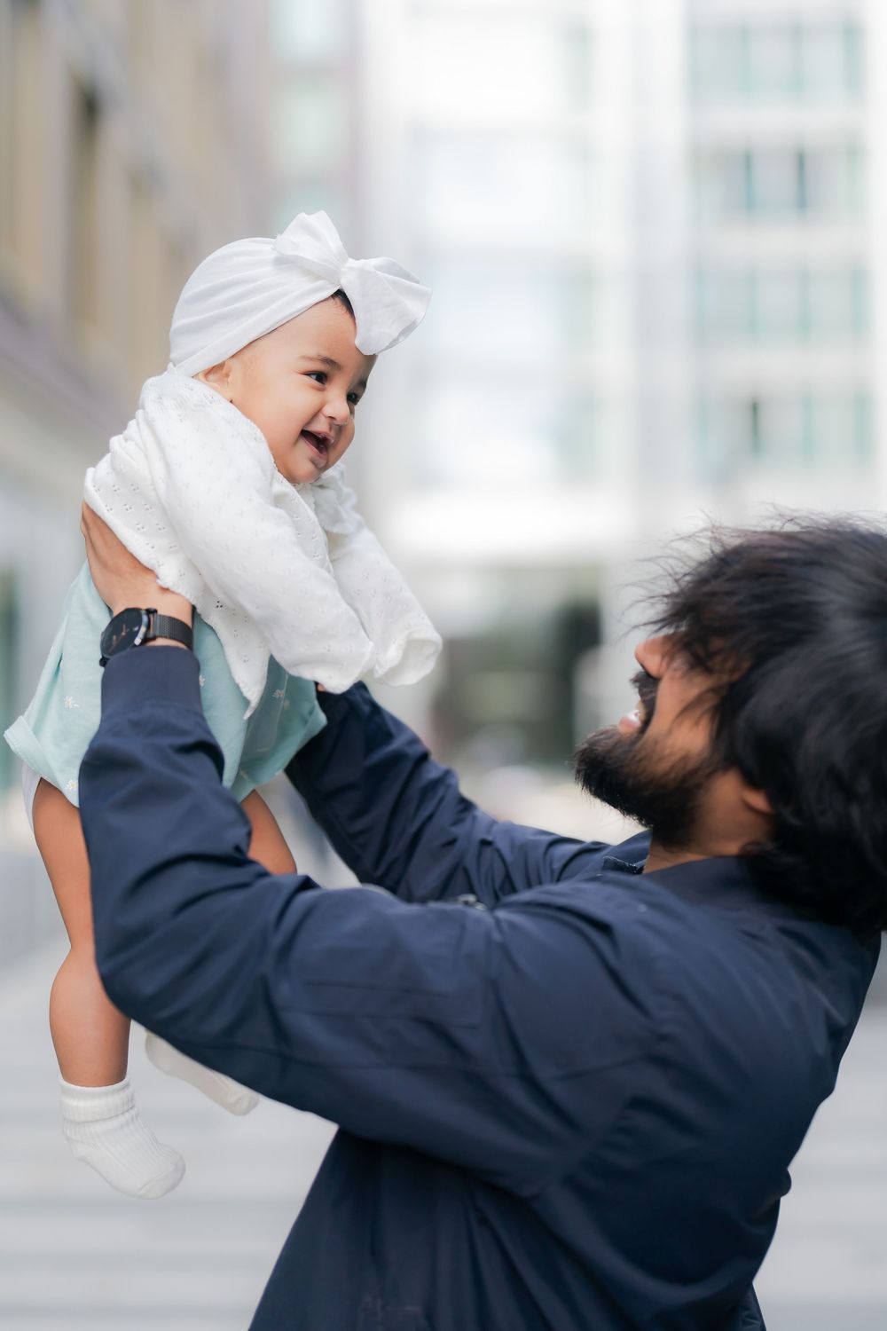 A bearded father joyfully lifts his laughing baby girl wearing a white bow turban outdoors in an urban setting.