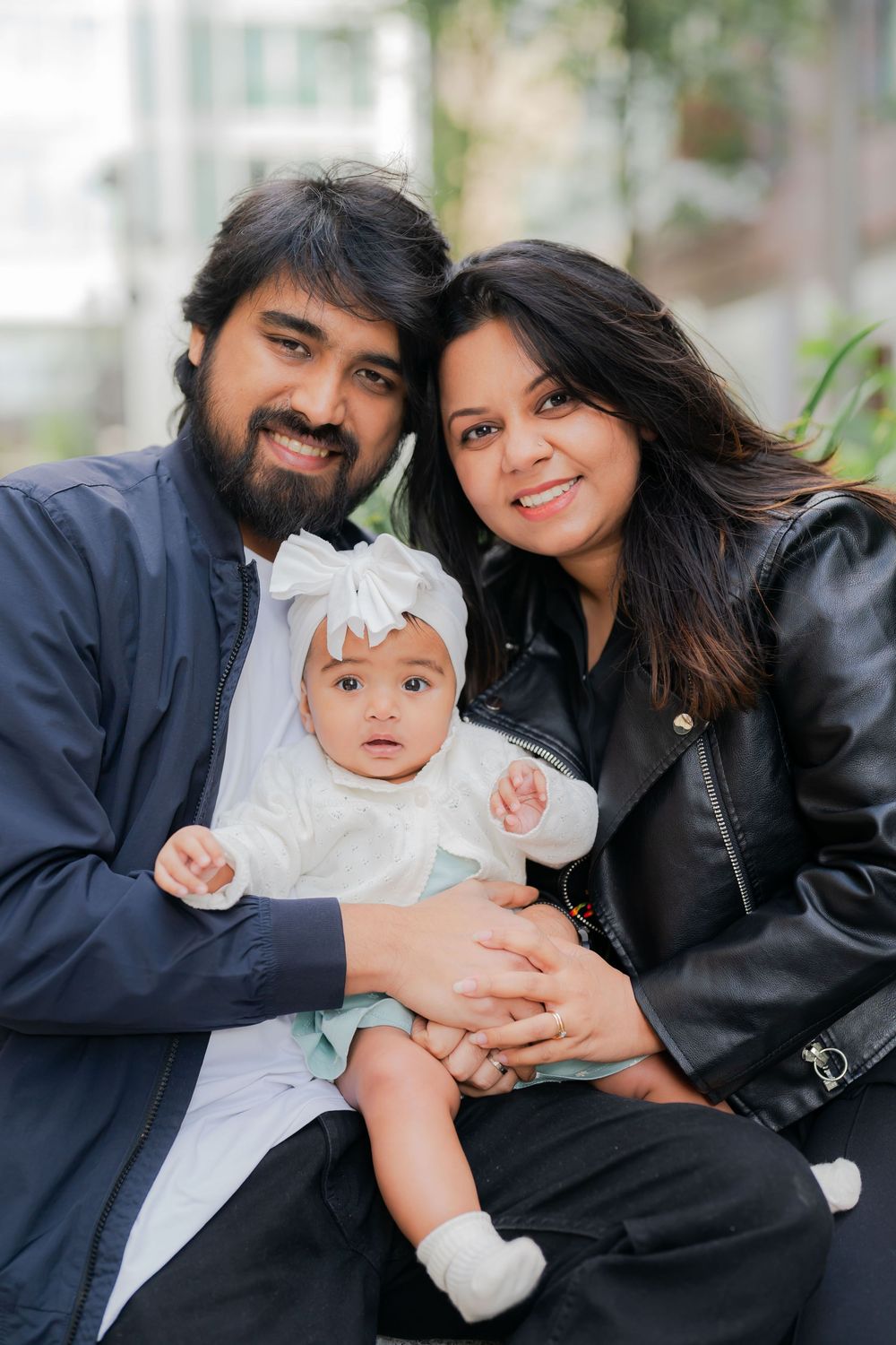 Happy family portrait with bearded father, smiling mother in leather jacket, and adorable baby girl in white bow headband.
