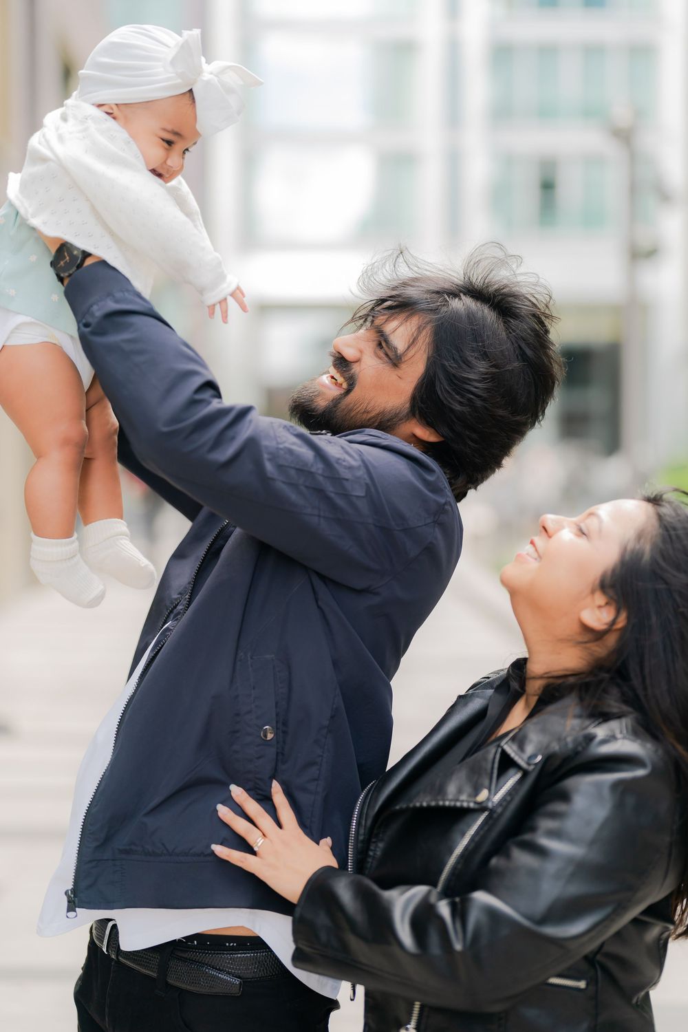 Happy family with father lifting baby girl in white outfit while mother looks on, outdoors in urban setting.