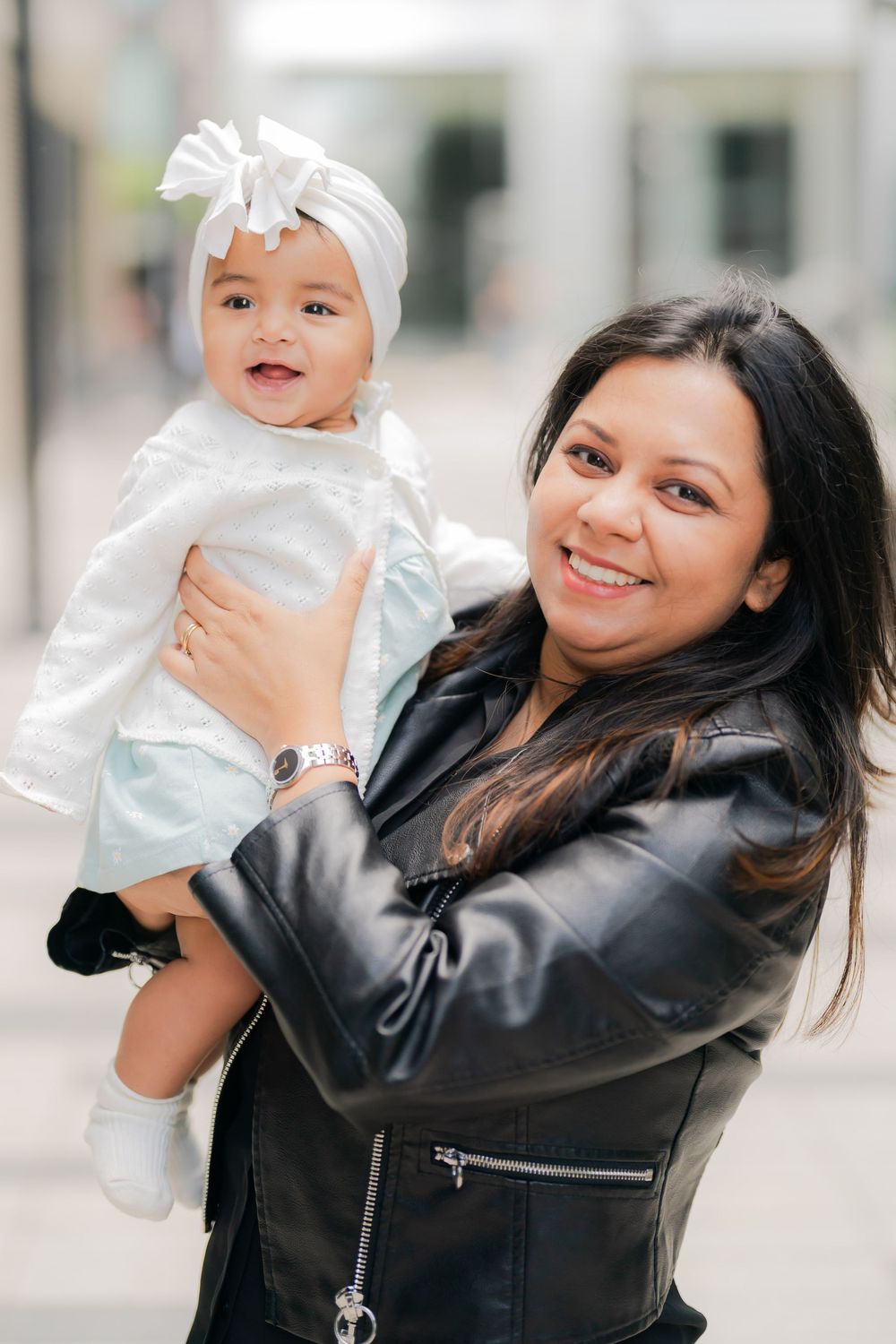 A smiling woman in a black leather jacket holds a happy baby dressed in white with a bow headband outdoors.