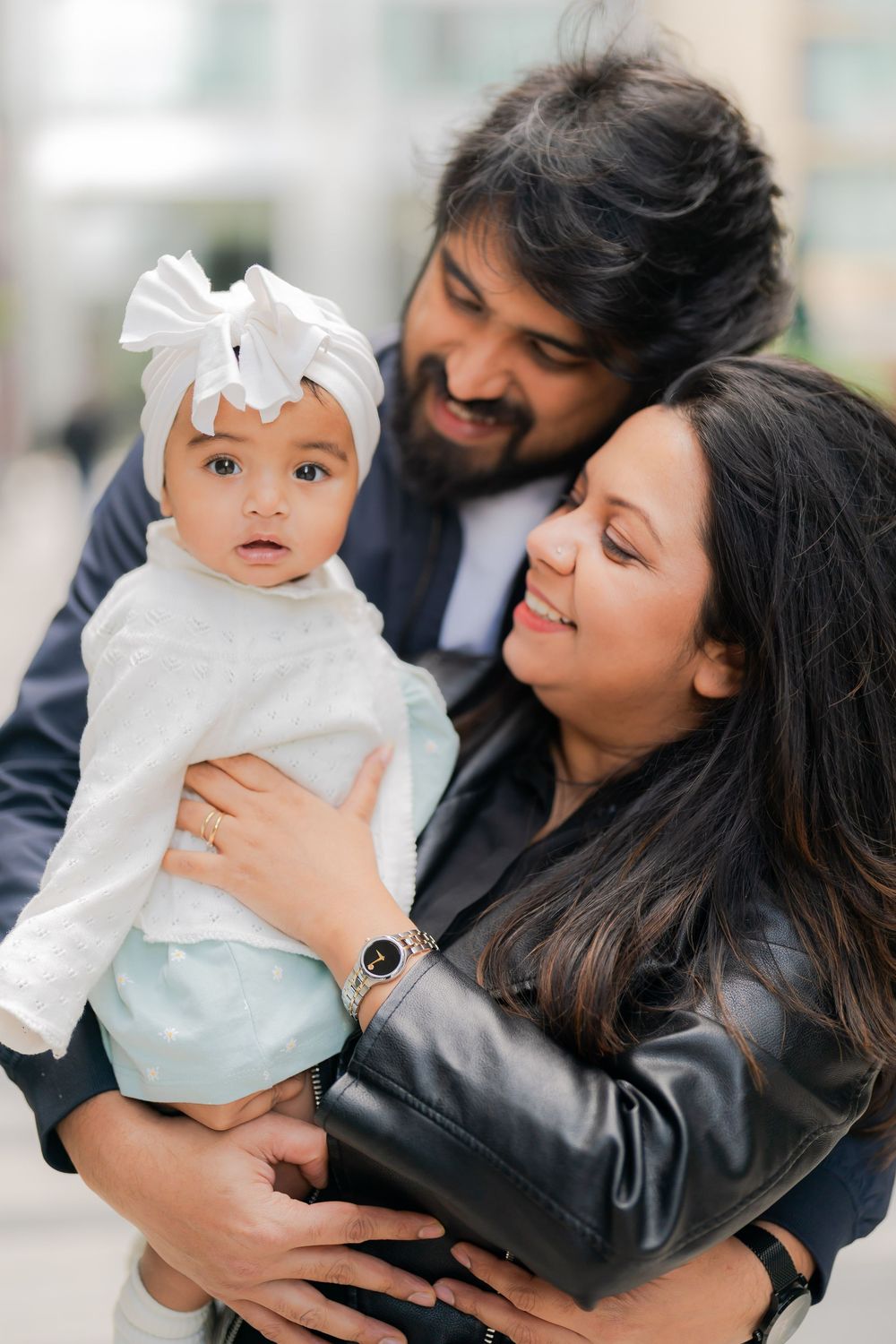 A smiling South Asian couple holds their baby girl dressed in white with a bow headband outdoors.