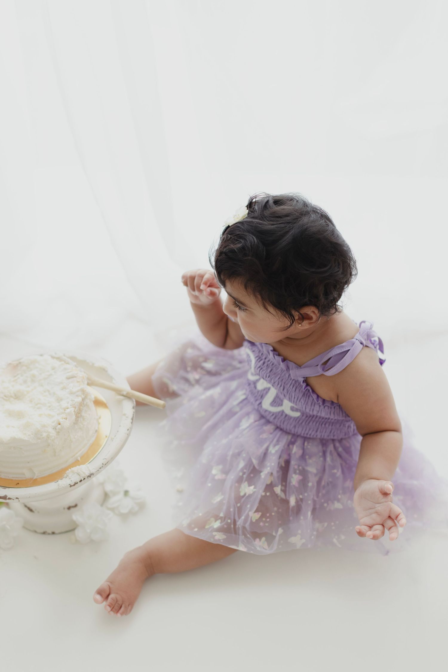 Baby girl in purple tutu dress sits beside white smash cake during first birthday photo session.