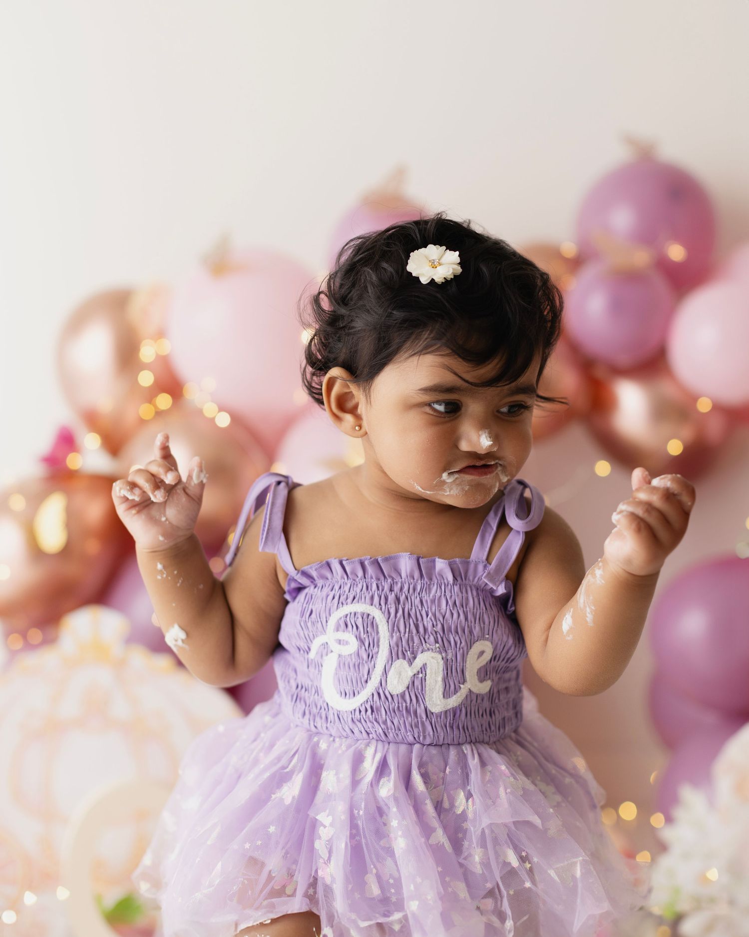 Baby girl in purple 'One' dress at first birthday cake smash with pink and rose gold balloons.