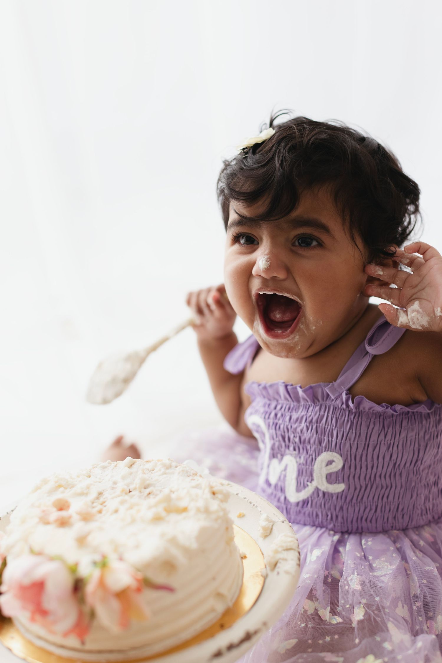 Excited toddler girl in purple dress celebrates first birthday smashing cake with spoon, face covered in frosting.