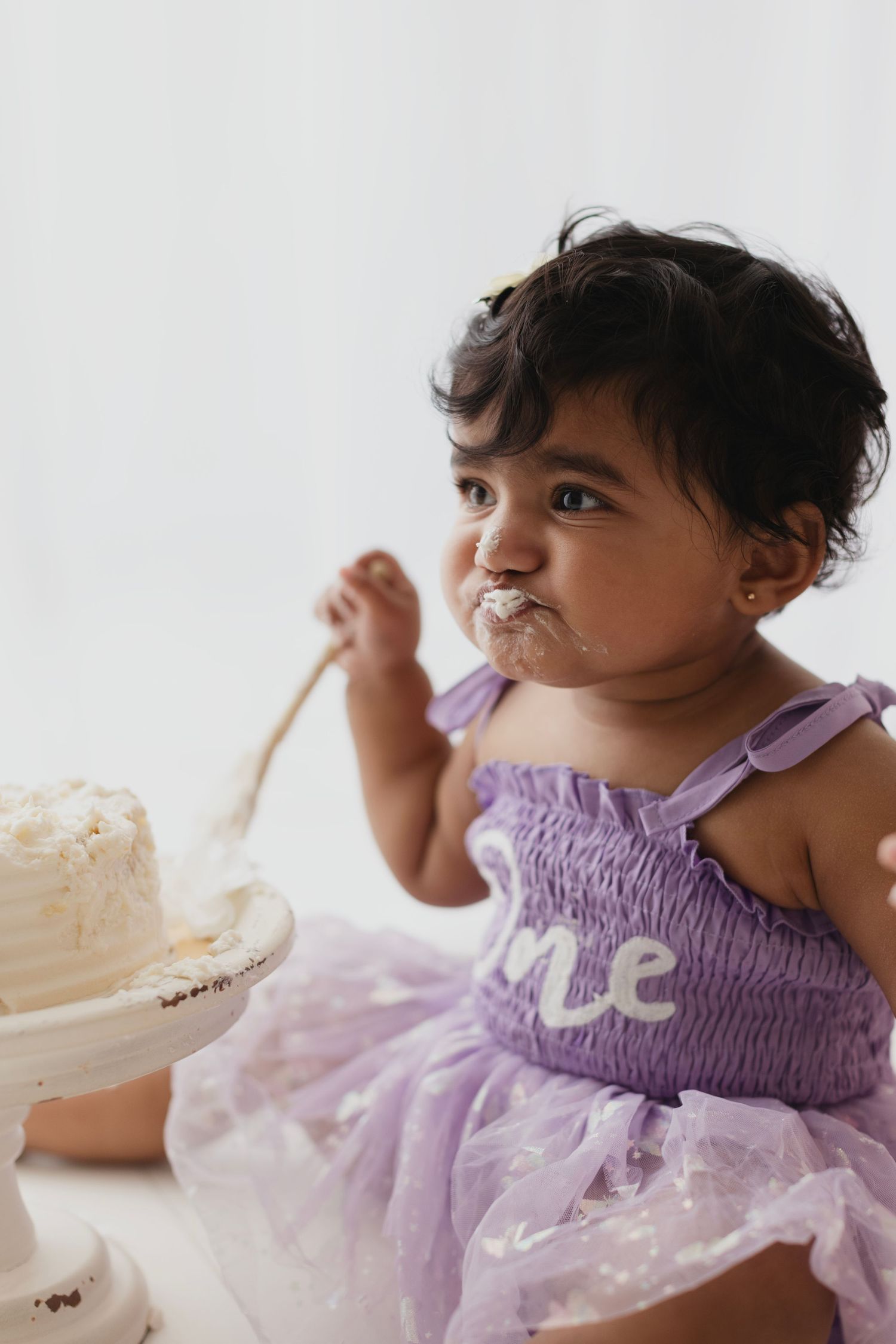 Baby girl in purple tutu dress eating first birthday cake with frosting on her face during cake smash photo session.
