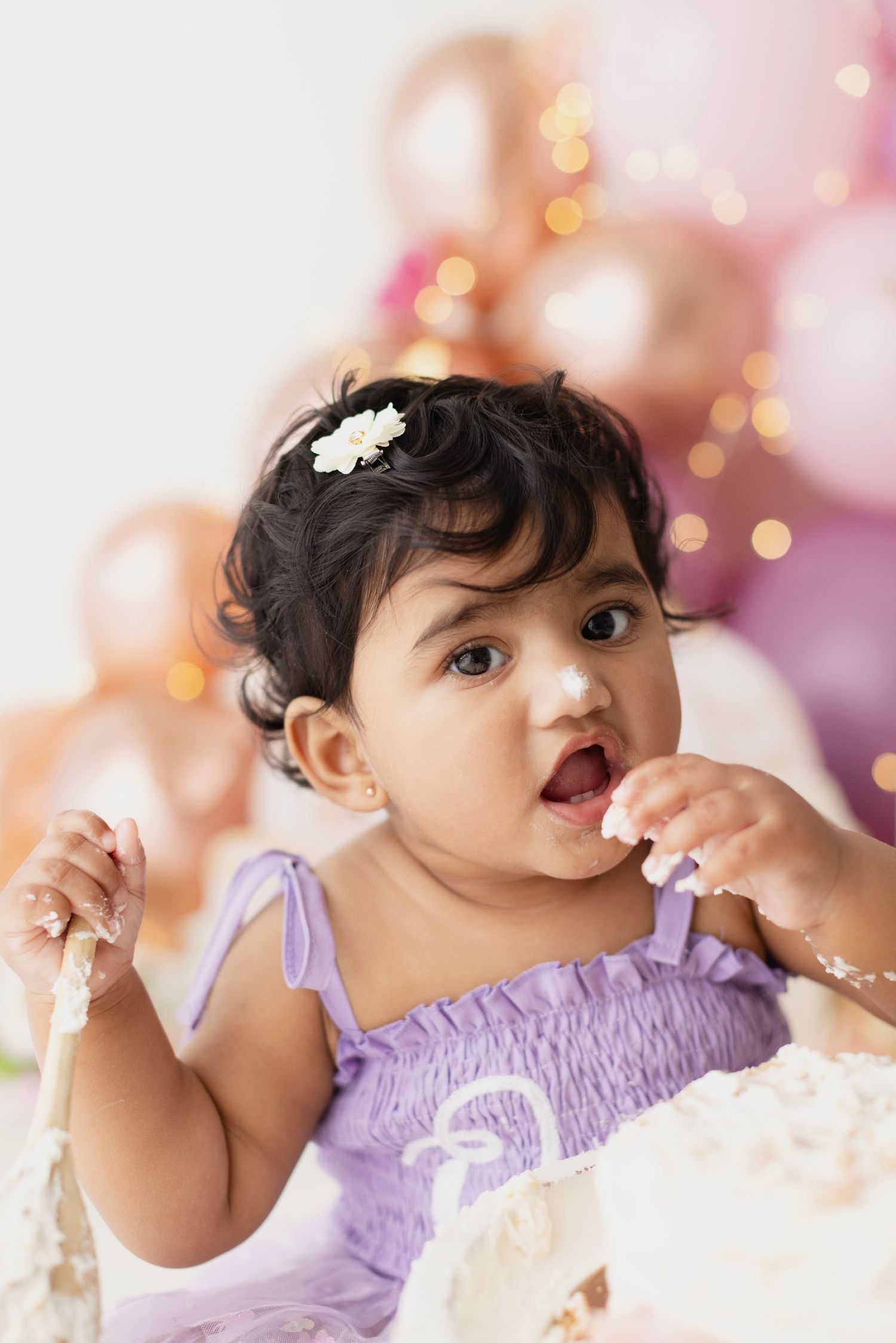 Baby girl in purple dress eating first birthday cake surrounded by pink and gold balloons.