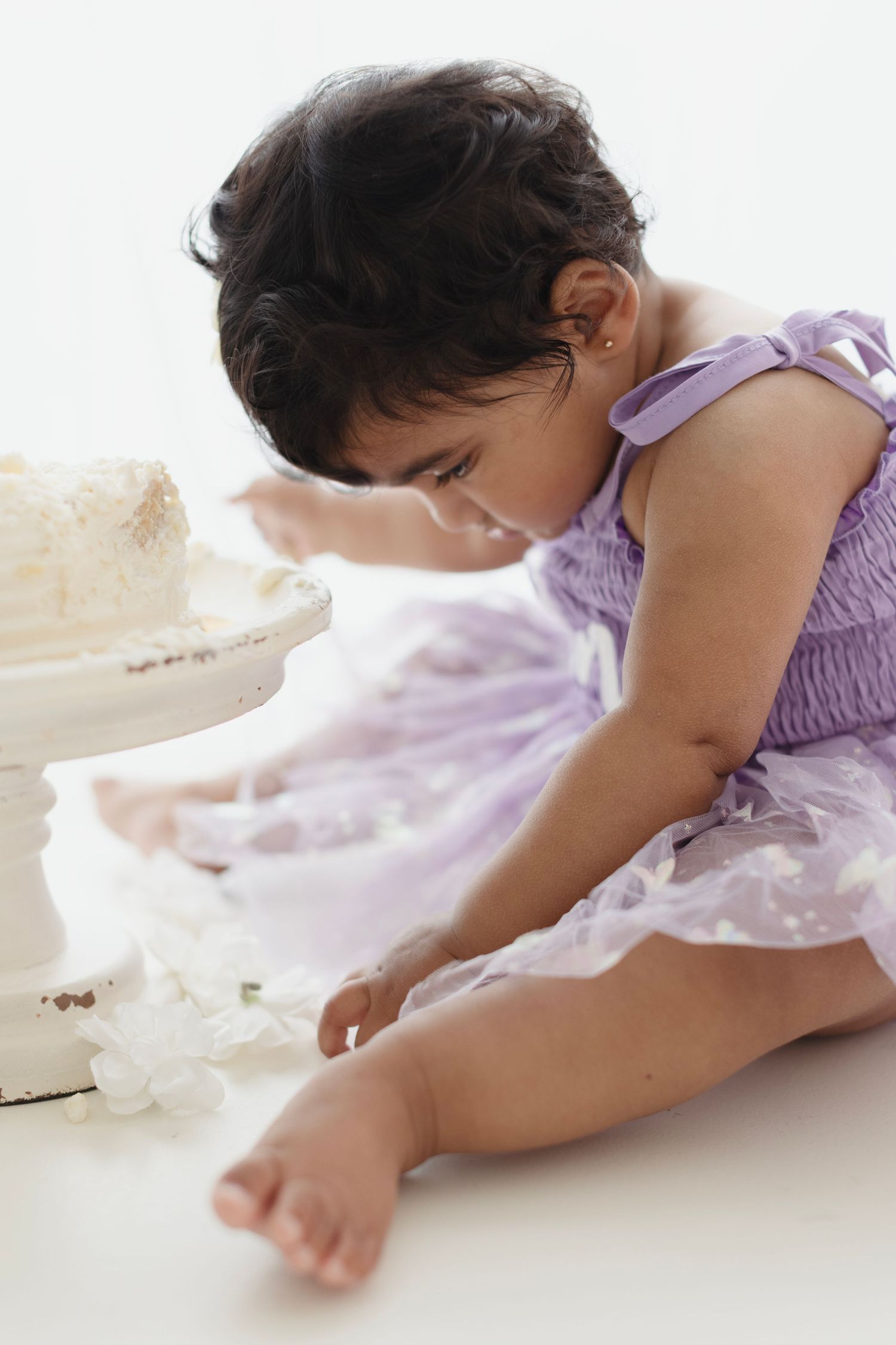 Baby girl in purple tutu dress reaching toward white cake during first birthday cake smash photo session.