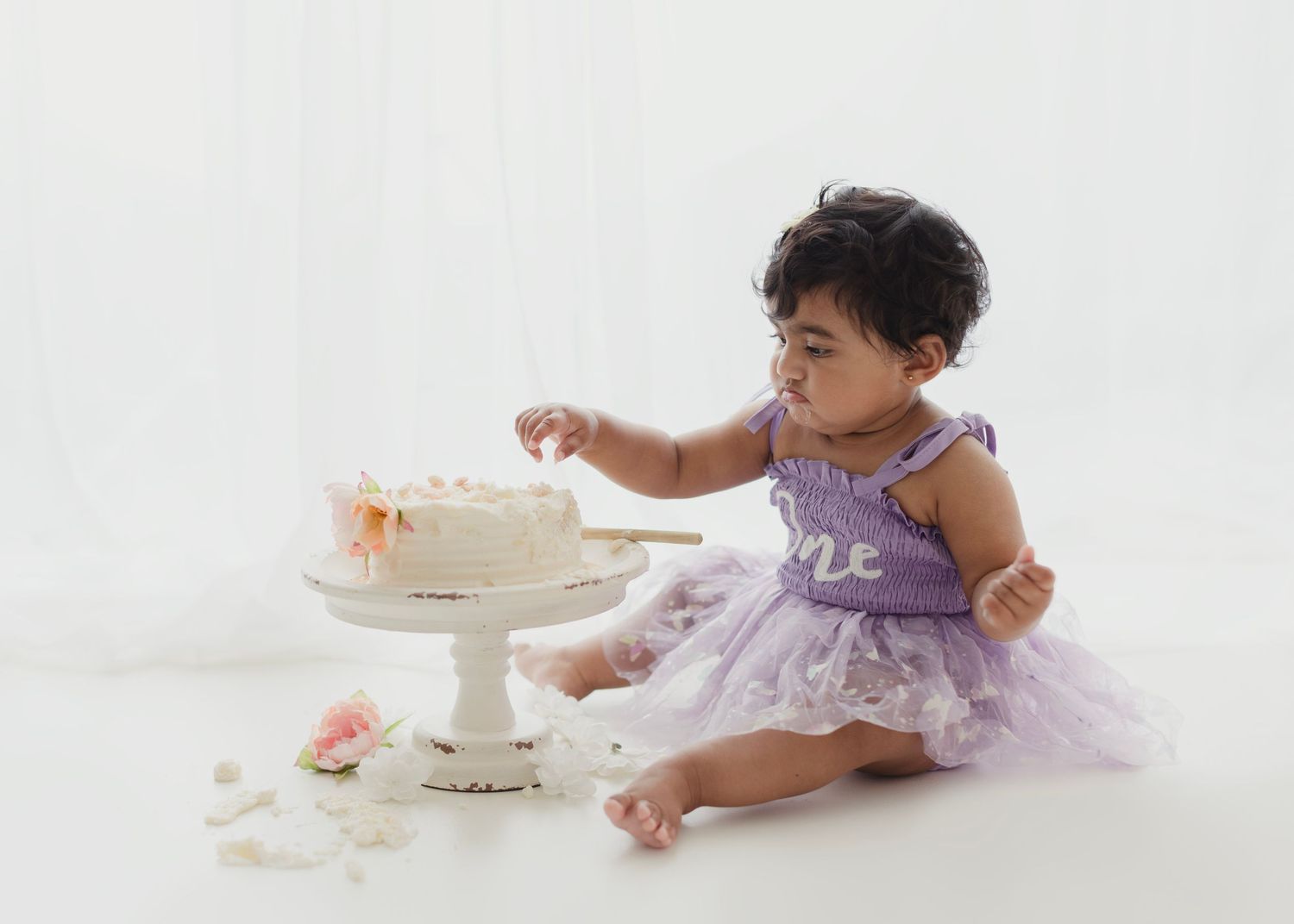 Baby girl in lavender tutu dress smashing her first birthday cake on white background.