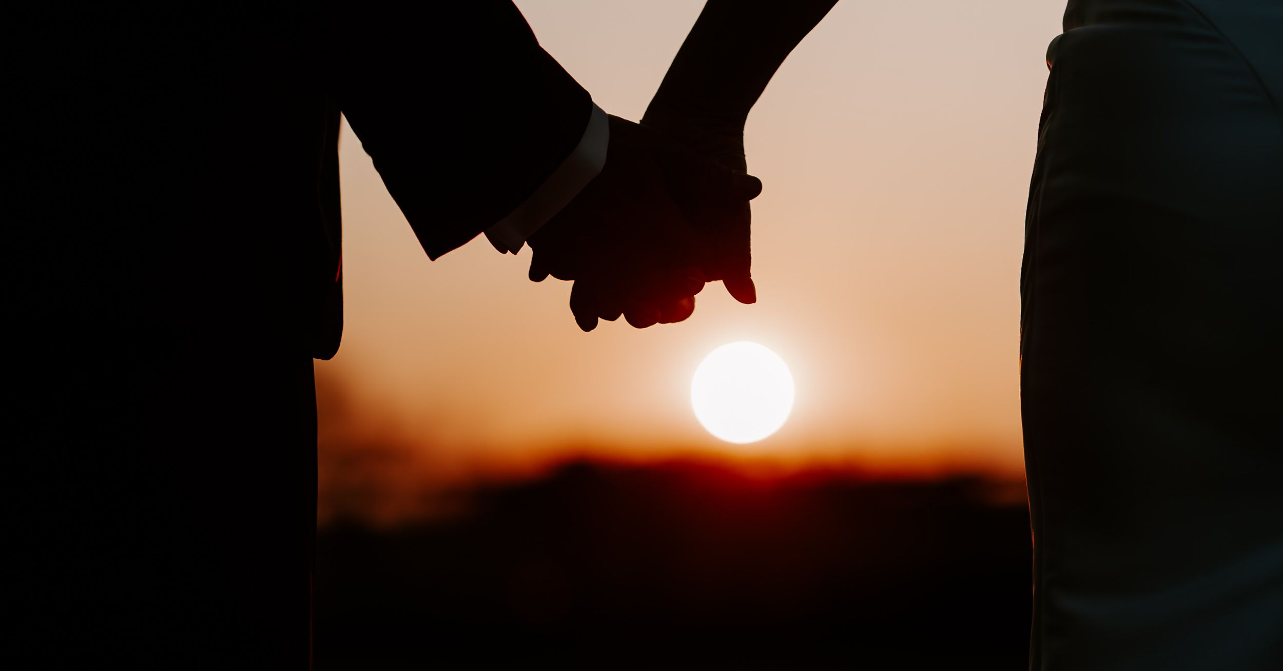 Silhouette of couple holding hands at sunset with warm orange sky backdrop.