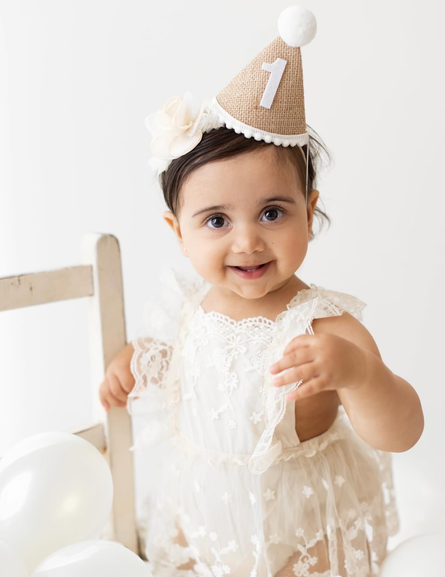 A smiling baby girl in a white lace dress wearing a burlap number one birthday hat celebrates her first birthday.