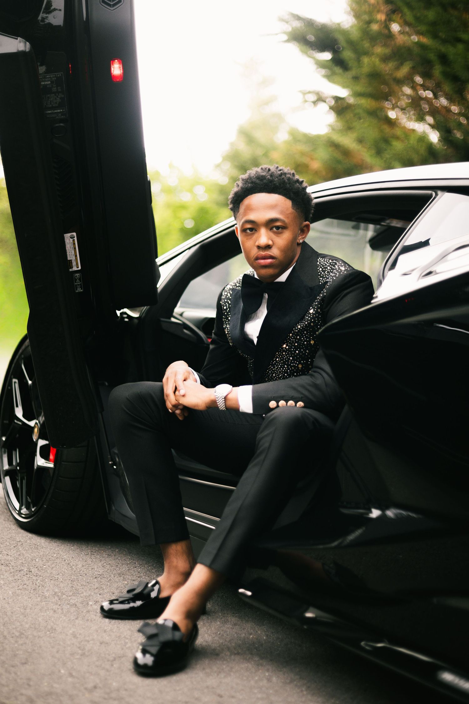 Young Black man in black embellished tuxedo sits in open door of luxury sports car.